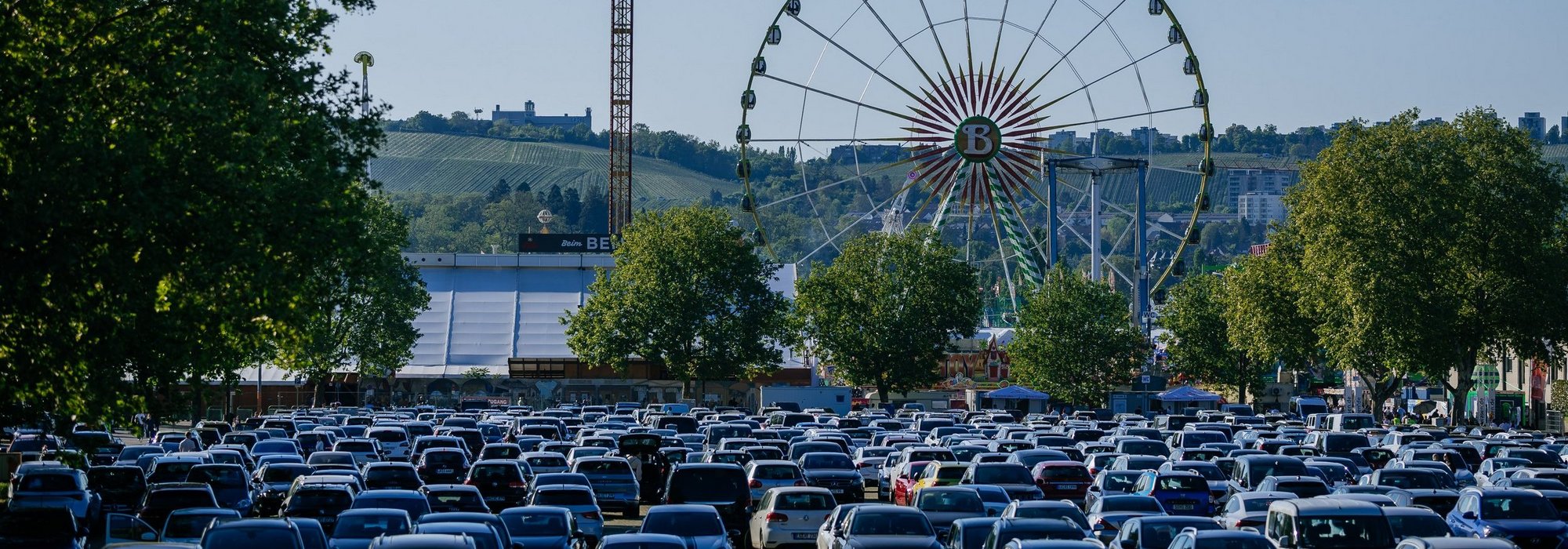 Unzählige Autos stehen auf einem Parkplatz. Im Hintergrund sieht man ein Riesenrad und ein Festzelt.