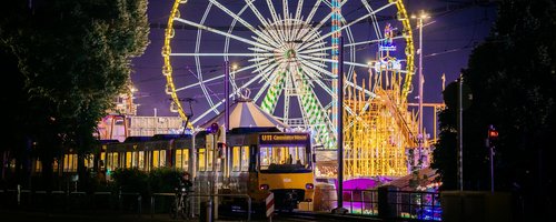 Eine Stadtbahn fährt in der Dunkelheit. Im Hintergrund sieht man das beleuchtete Riesenrad und andere Fahrgeschäfte.
