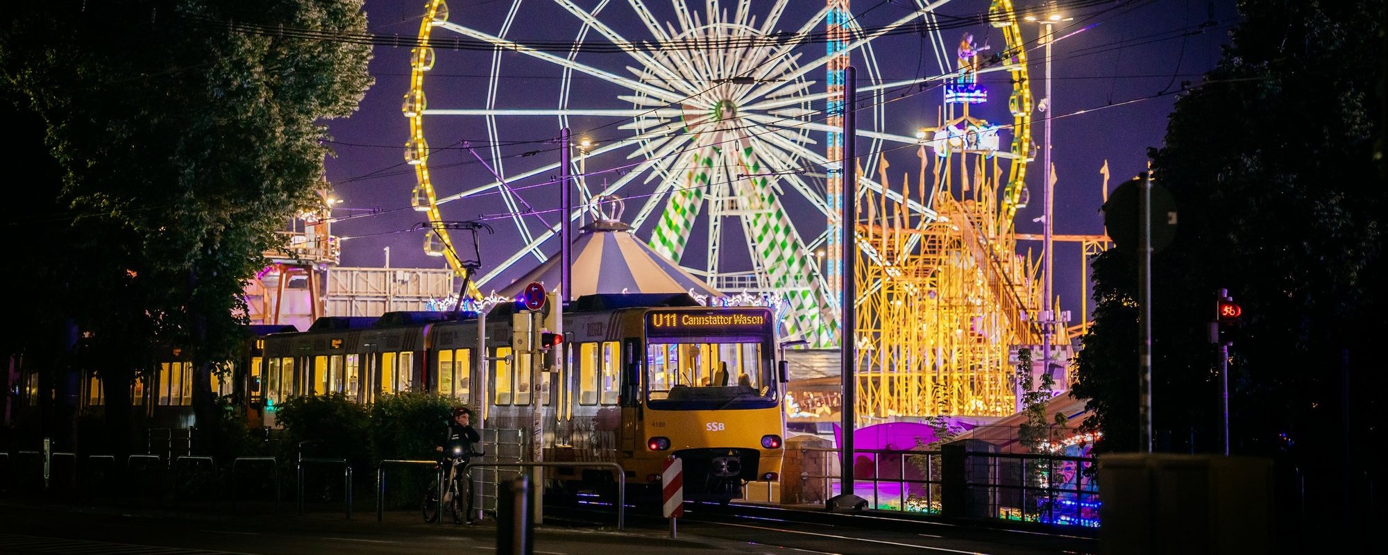 Eine Stadtbahn fährt in der Dunkelheit. Im Hintergrund sieht man das beleuchtete Riesenrad und andere Fahrgeschäfte.