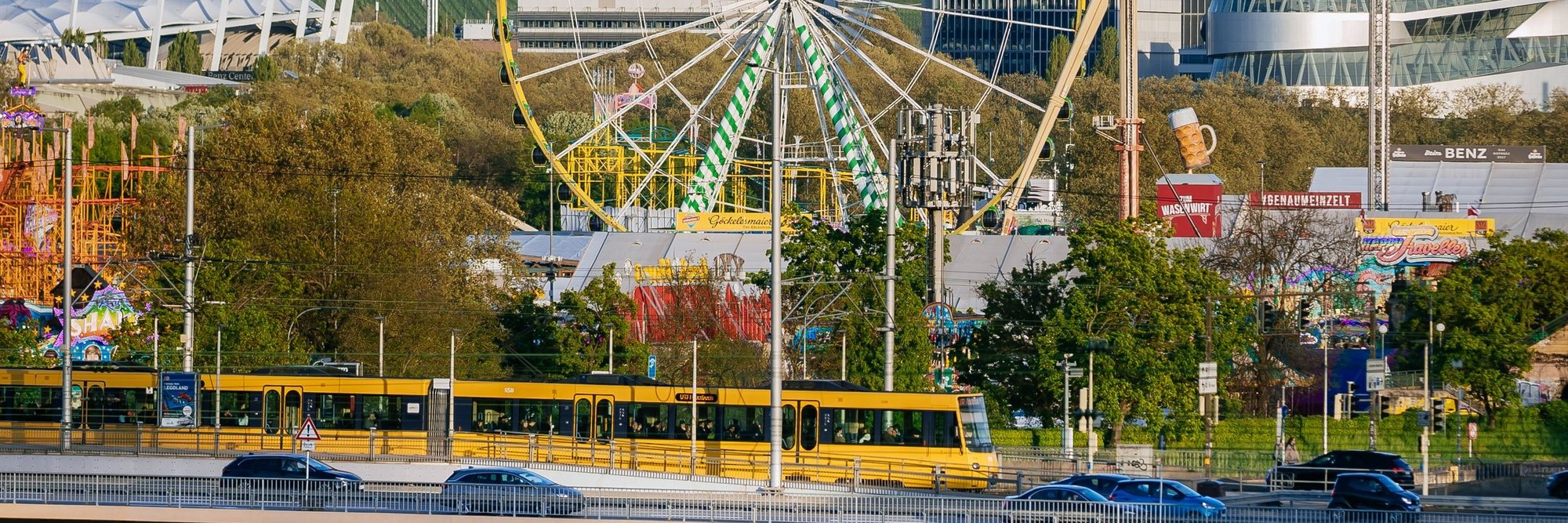 Blick auf den Neckarpark. Im Vordergrund fährt eine Stadtbahn vorbei. Dahinter sind die Fahrgeschäfte des Frühlingsfestes, die MHPArena und das Mercedes-Benz-Museum erkennbar.