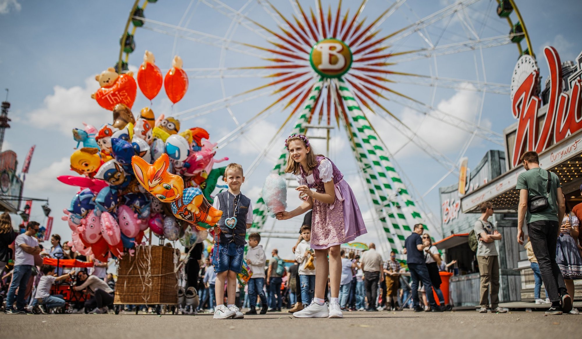 Ein Junge und ein Mädchen stehen auf dem Volksfestplatz mit Zuckerwatte in der Hand. Hinter ihnen steht ein Luftballon-Verkäufer. Dahinter steht das Riesenrad.