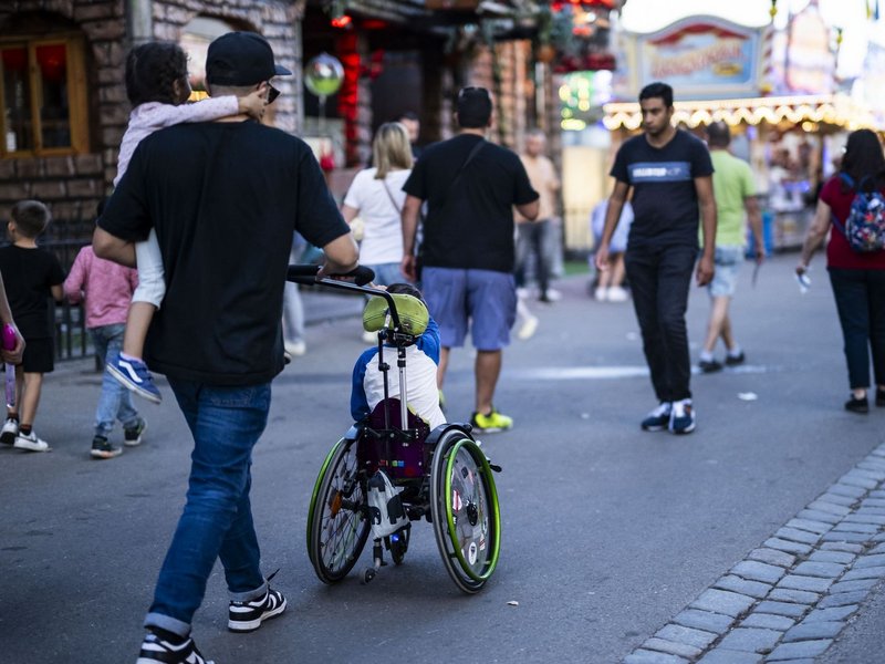 Ein Mann schiebt ein Kind im Rollstuhl über den Wasen und hält gleichzeitig noch ein Kind auf dem Arm.
