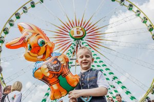 Ein Junge steht mit einem Luftballon von Wasen-Maskottchen Wasi vor dem Riesenrad Bellevue.