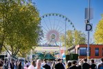Blick auf Wasen-Eingangstor mit Schriftzug "Herzlich willkommen!". Im Vordergrund Menschen. Im Hintergrund Riesenrad und blauer Himmel.