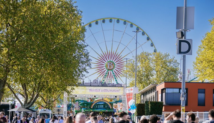 Blick auf Wasen-Eingangstor mit Schriftzug "Herzlich willkommen!". Im Vordergrund Menschen. Im Hintergrund Riesenrad und blauer Himmel.