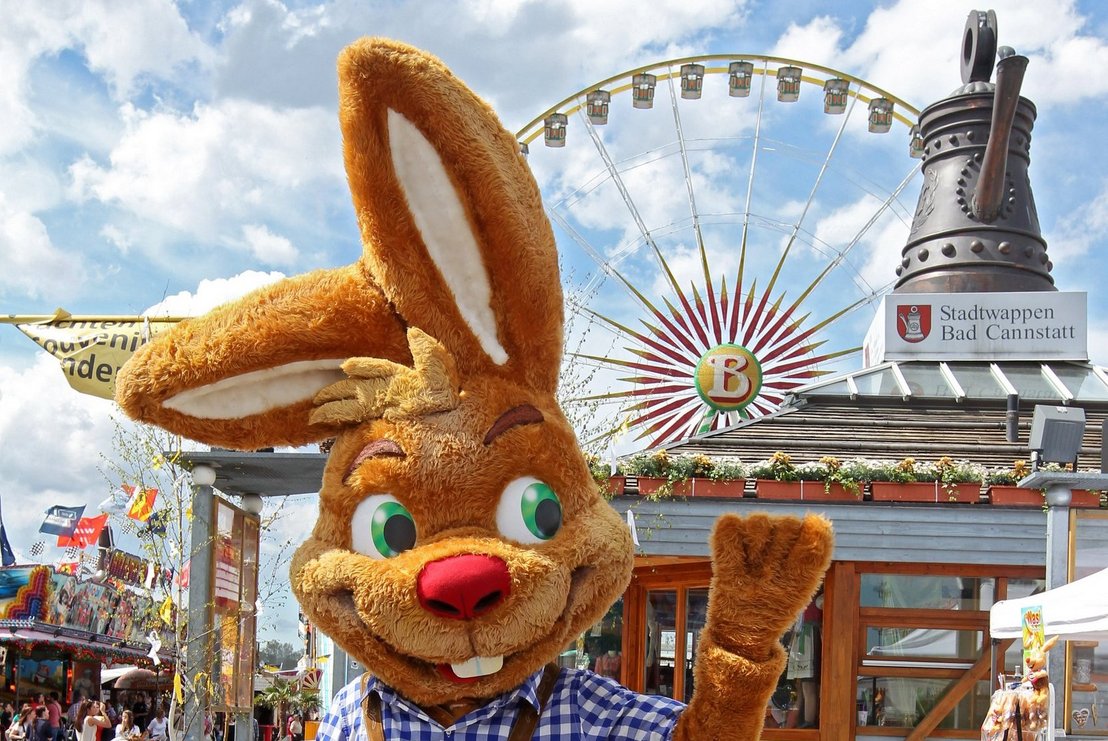 Das Wasen-Maskottchen Wasi winkt fröhlich vor dem Info-Pavillon mit der Cannstatter Kanne auf dem Dach.