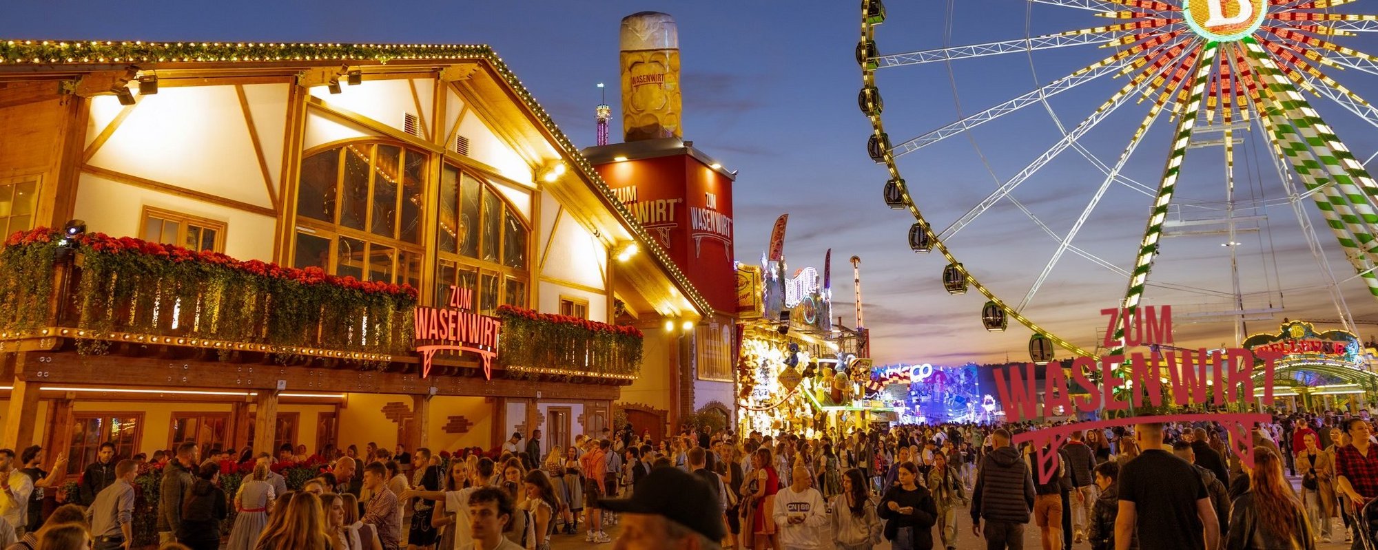 Aussenansicht des Wasenwirt-Festzeltes in der Dämmerung mit Beleuchtung. Daneben steht das leuchtende Riesenrad.