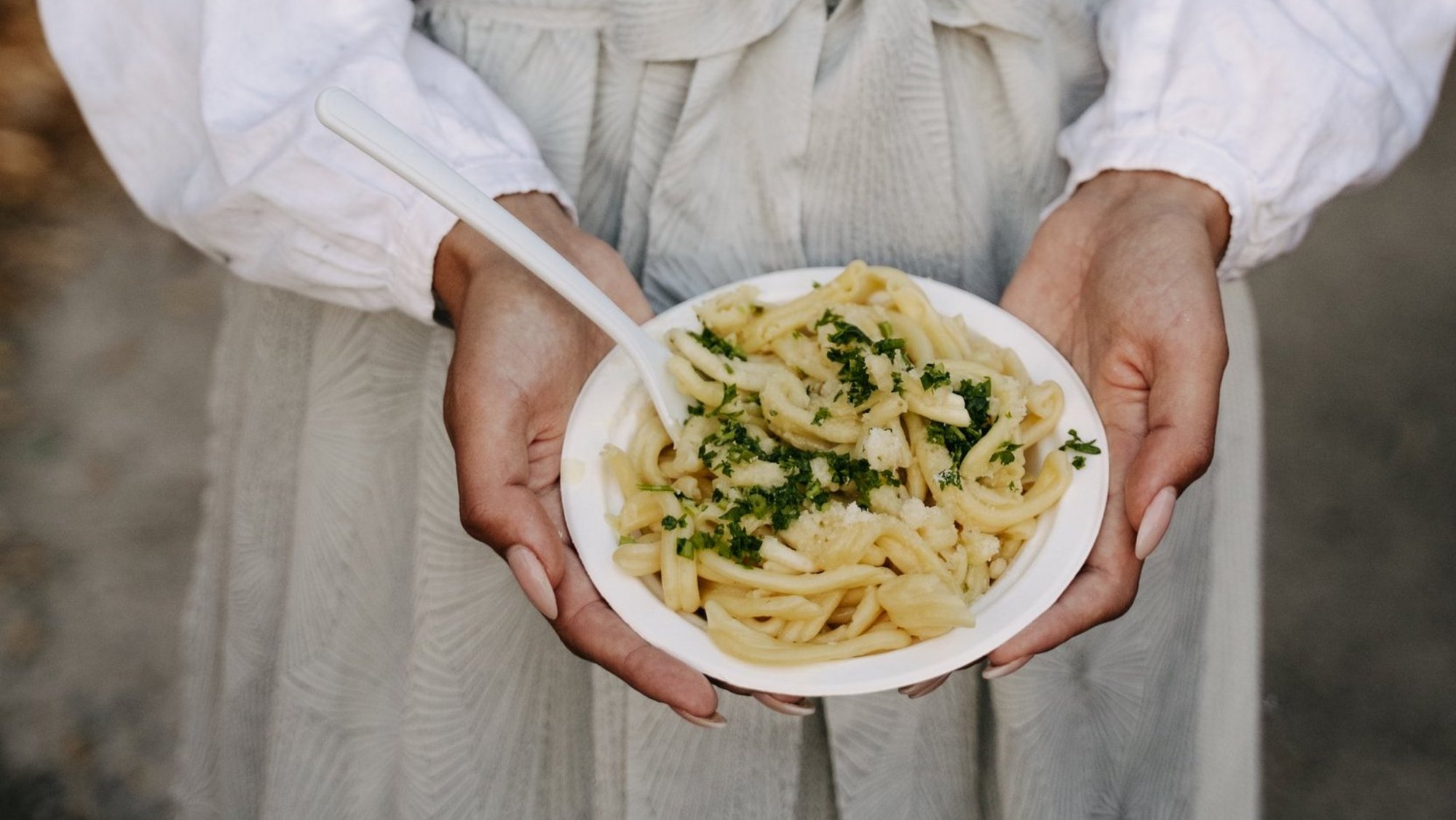 Eine Frau in einem grauen Dirndl hält einen Teller mit Schupfnudeln in die Kamera.