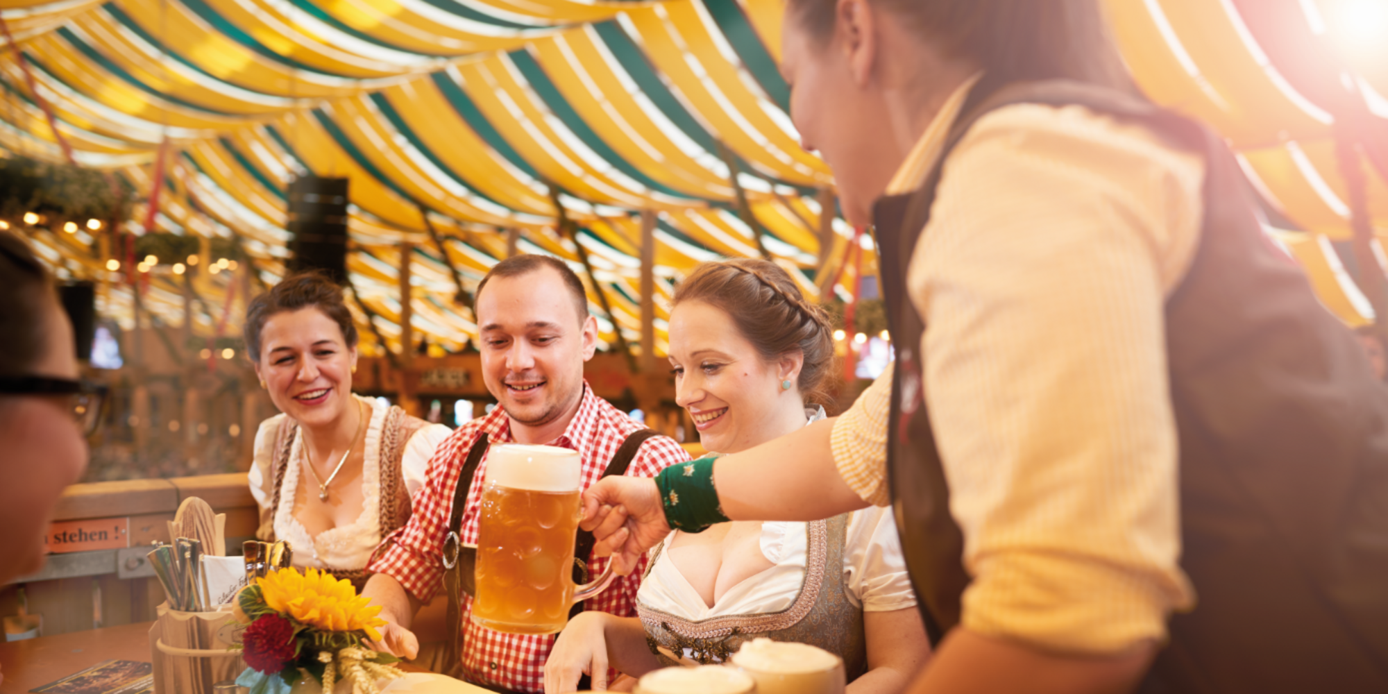Zwei Frauen und ein Mann sitzen an einem Biertisch im Zelt. Ein Kellner stellt einen Maßkrug auf den Tisch.