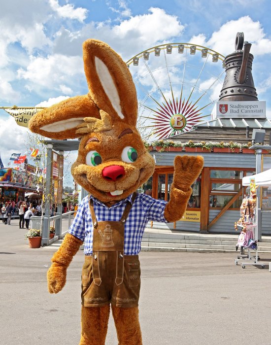 Maskottchen "Wasi" steht vor dem Info-Pavillon auf dem Cannstatter Wasen und winkt fröhlich in die Kamera.