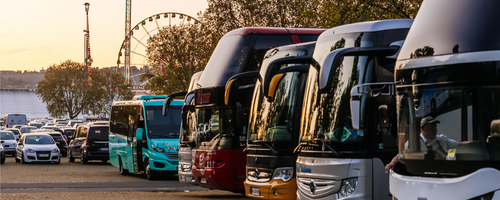 Mehrere Reisebusse stehen auf einem Parkplatz. Im Hintergrund sieht man die Fahrgeschäfte des Frühlingsfestes.