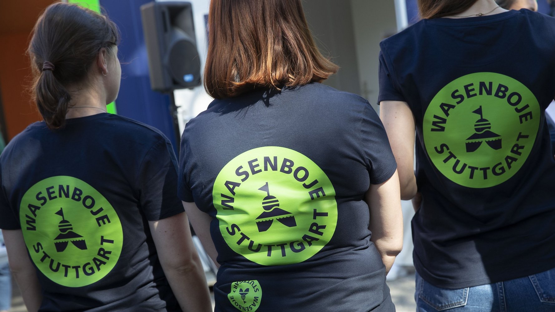 Drei Frauen stehen mit dem Rücken zum Fotografen und haben auf ihren dunklen T-Shirts einen grün leuchtenden Kreis aufgedruckt, in dem "Wasenboje Stuttgart" steht.