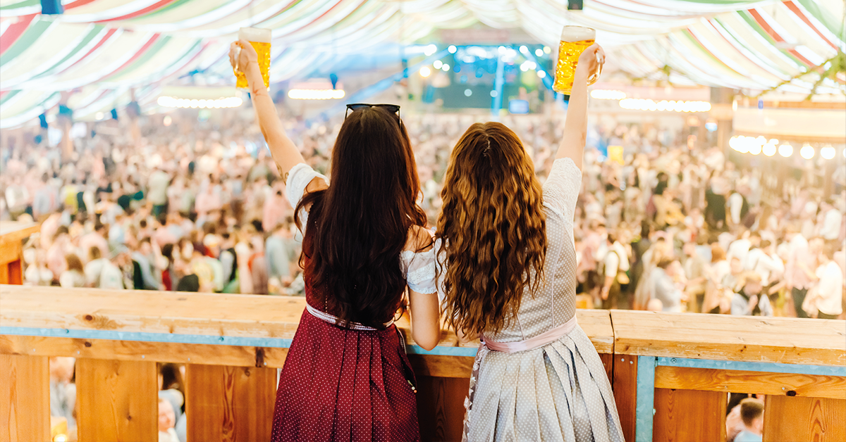 Zwei Frauen in Tracht heben ihre Maßkrüge auf einem Balkon nach oben und prosten der feiernden Menge unter ihnen zu.